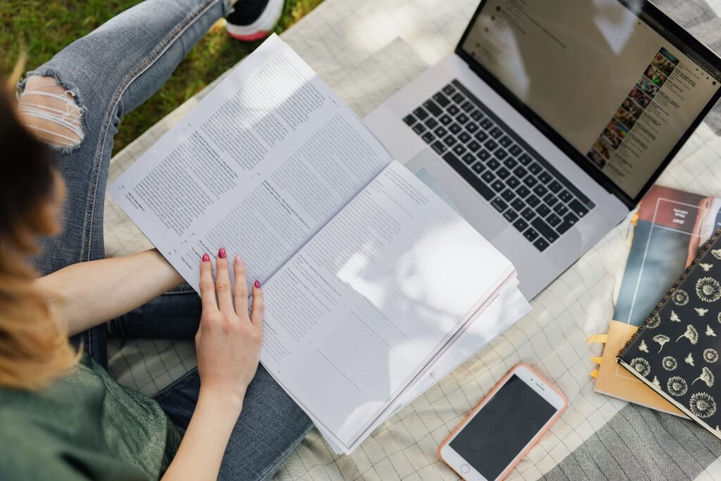 High angle of crop anonymous female student studying information in textbook while sitting with modern laptop and smartphone in sunny park
