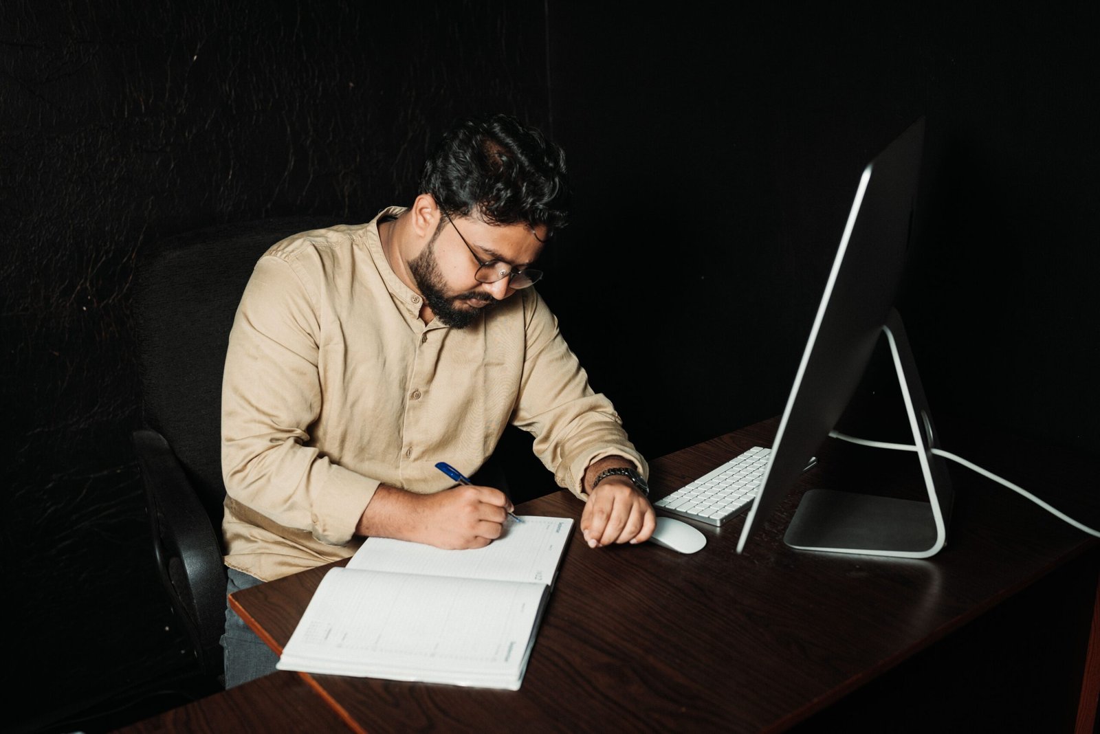 Man writing in office with computer. Late-night work focused on creativity and productivity.