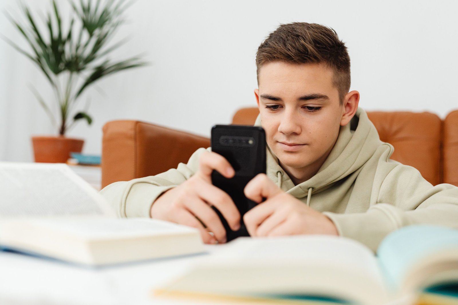 Focused teenager in a hoodie using a smartphone surrounded by books, indoors.