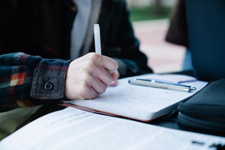 Close-up of an individual writing in a notebook outdoors, featuring pens and paper.