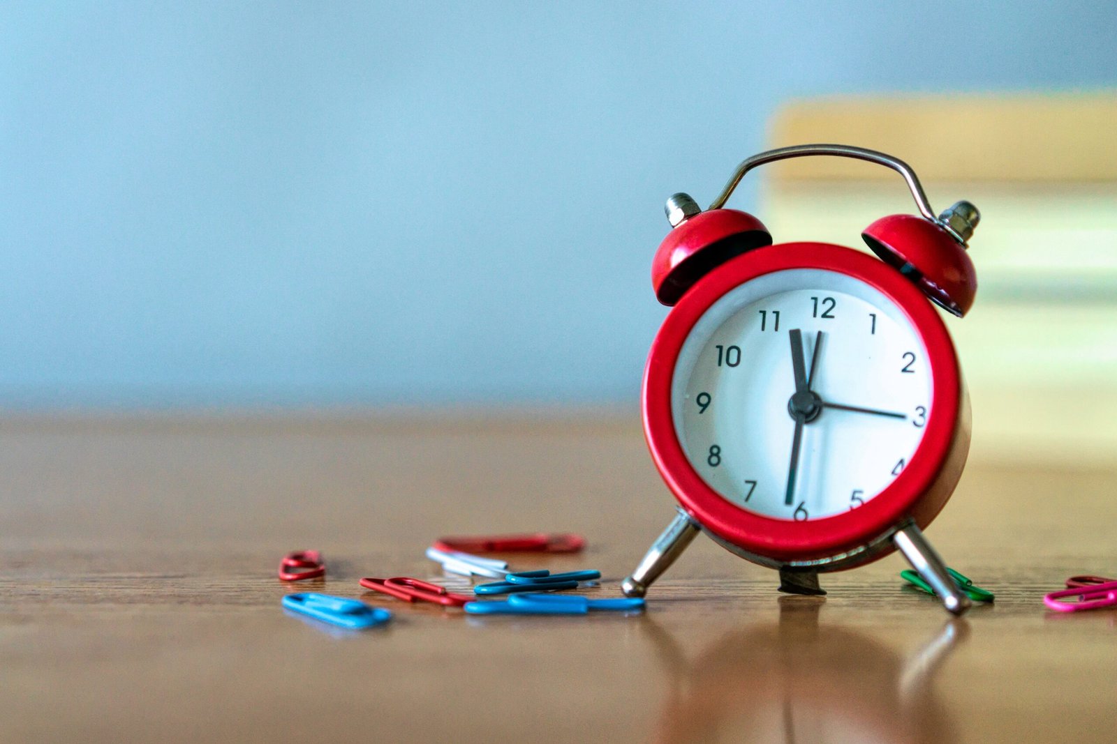 Red alarm clock and colorful paper clips on a wooden desk in a close-up view.